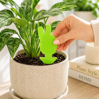 Person holding a green cactus-shaped plant marker next to a potted plant on a wooden surface.